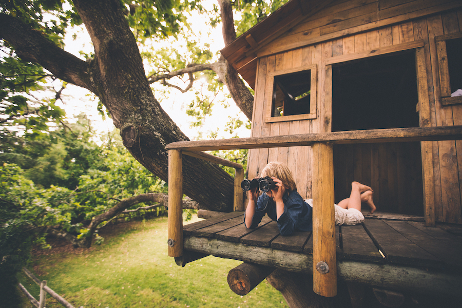 Young boy lying on the porch of a treehouse using binoculars to spy on something in the distance with leaves and a lush green grassy park in the background